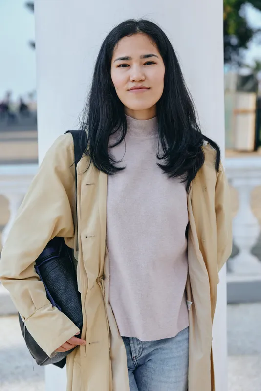 Woman wearing beige trench coat and soft pink sweater standing outdoors with jeans and bag, casual street style, confident expression, daytime light in urban setting