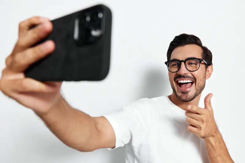 Happy smiling man in glasses takes a selfie with smartphone, showing thumbs up while standing against a light plain background. Social media lifestyle concept.