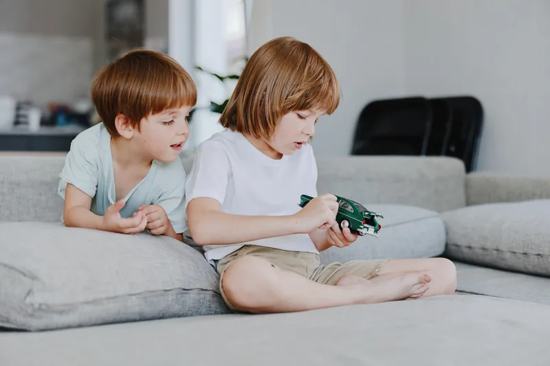 Two happy boys playing together with a toy car in a bright, modern living room. Their facial expressions reflect joy and curiosity as they engage in playtime.