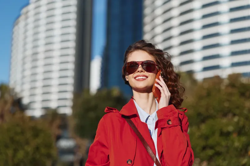 Confident young woman in a red trench coat smiles while talking on her phone against a modern city skyline with glass towers and bright sunlight in the background.