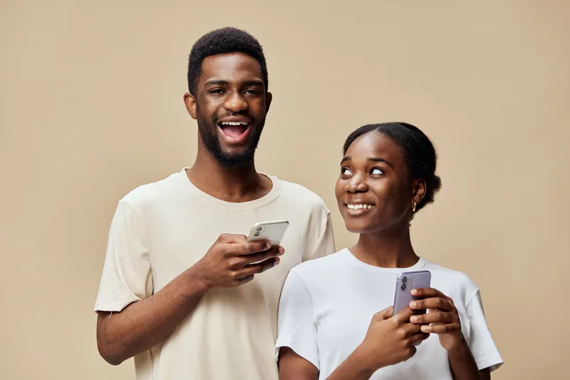 Happy young African couple using smartphones together against a beige background, showcasing joy and connection through technology