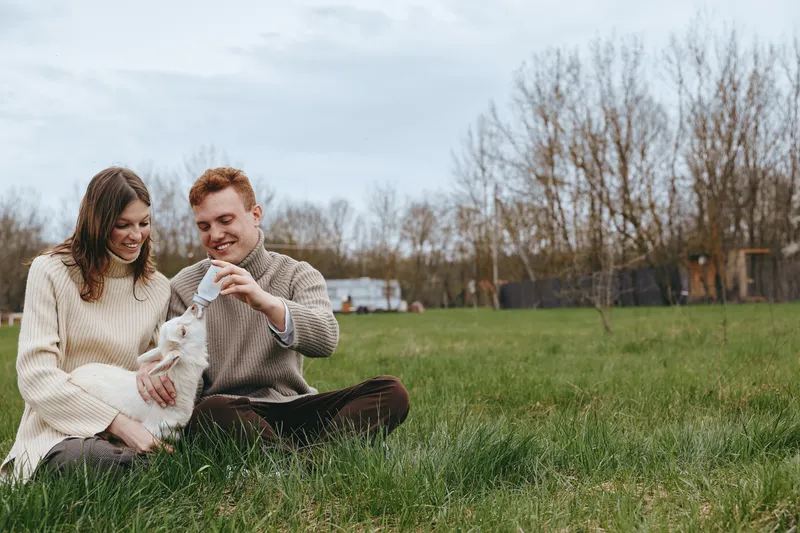 Happy couple outdoors on a grassy field softly feeds two kid goats, sharing smiles and a cozy moment, casual sweaters, warm light, relaxed lifestyle shot, natural colors, affectionate animal