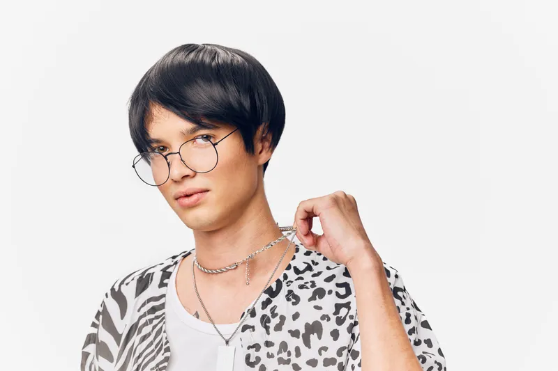 Young man with glasses wearing patterned shirt and silver chain posing confidently against white background, showing modern casual style and subtle expression.