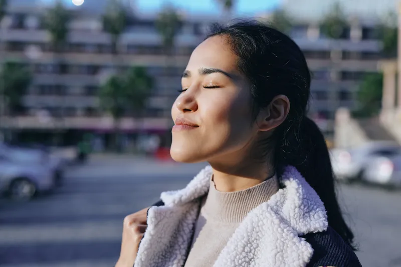 Woman with closed eyes enjoying sunlight outdoors, wearing a fleece jacket, calm moment in a warm breeze, peaceful mood, natural light on her face, relaxed