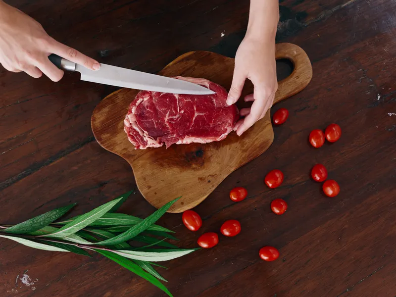 Женщина с ножом в руке нарезает свежее мясо стейка для жарки на кухне, на столе соль, перец и другие специи, красные помидоры черри и зелень, приготовление ужина