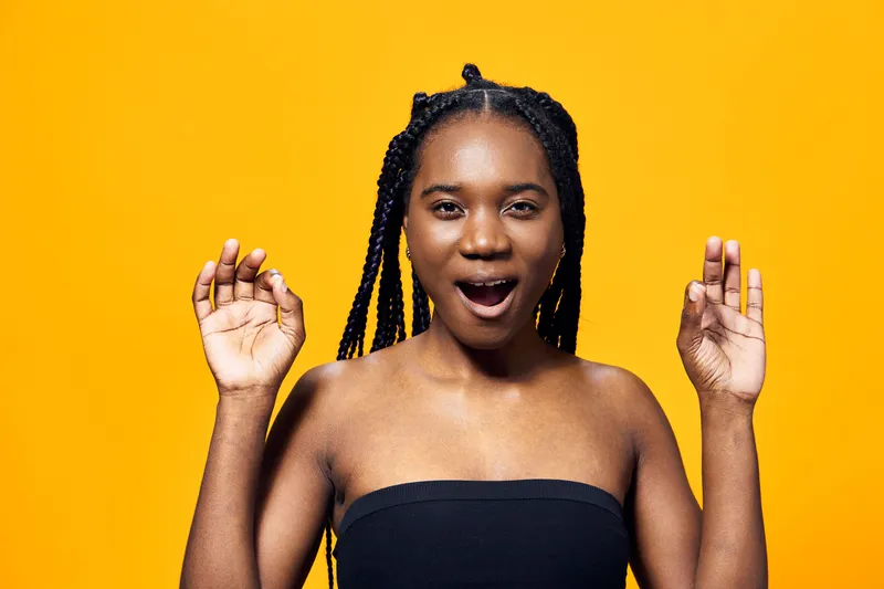 Young Black woman with braids expressing joy against a vibrant yellow background, highlighting happiness, fashion, and enthusiasm in a summer inspired setting