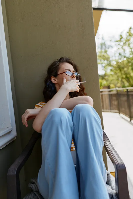 Woman sits on a balcony wall outdoors, smoking a cigarette, relaxed casual style, wearing light blue pants and glasses, natural daylight candid lifestyle scene, urban vibe