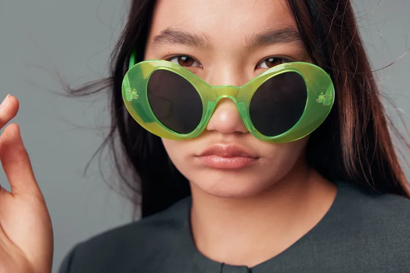 Woman with long dark hair wearing large green sunglasses and dark clothing, close-up portrait with neutral expression on gray background in studio for fashion or style concept.