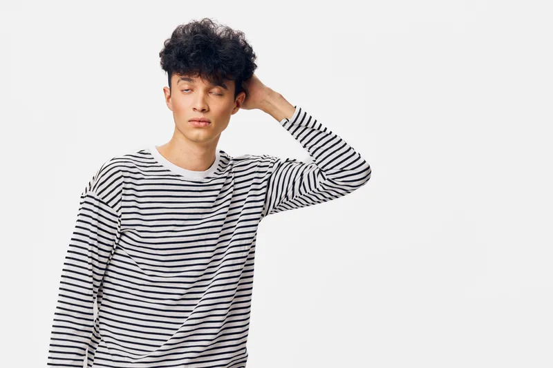 Young man in striped shirt with curly hair standing against white background, posing with thoughtful expression and hand on head, casual fashion style.
