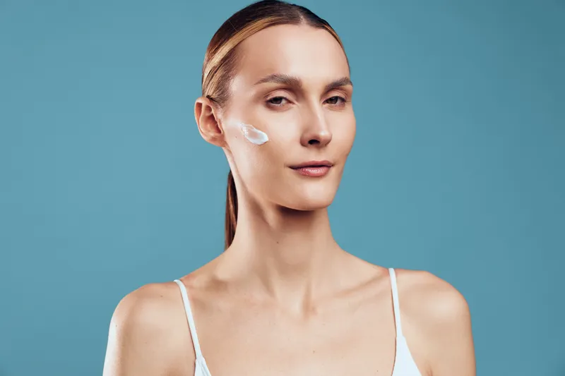 Young blonde woman applies a dollop of facial cream on her cheek in a studio with a blue backdrop, conveying a calm skincare moment and focused self care for a fashion of clean beauty and everyday