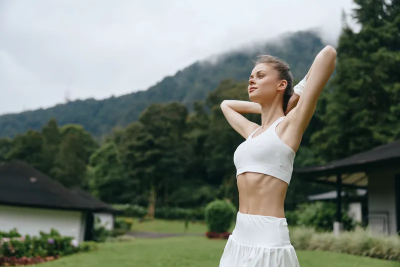 Young athletic woman in a white crop top and skirt enjoying nature, embodying a healthy lifestyle in a tranquil outdoor setting with mountains