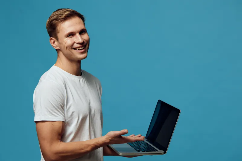 Smiling man holding open laptop computer while looking at camera, blue background, casual clothing, positive expression, technology concept, studio portrait, lifestyle image.