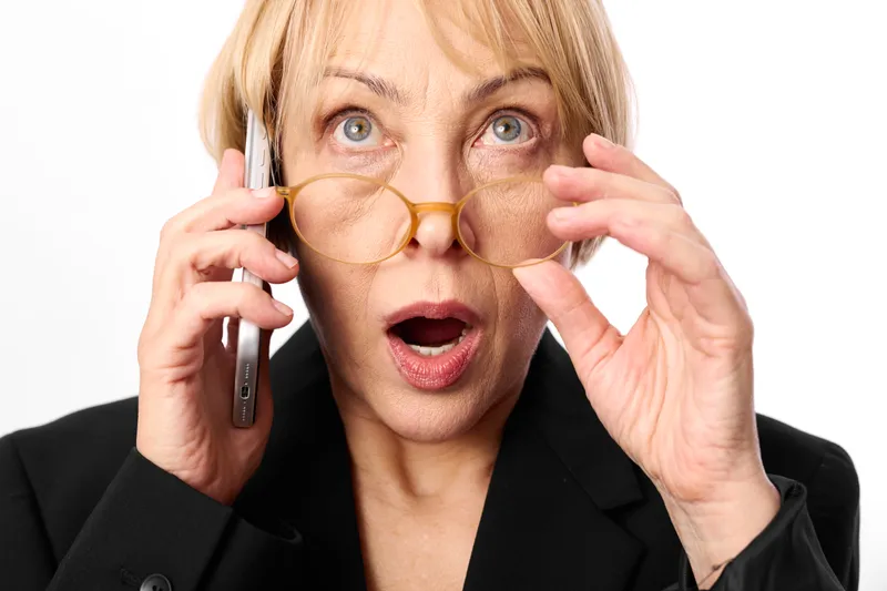 Surprised businesswoman with glasses holding phone, shocked eyes and open mouth, black blazer, white office background, urgent communication for stress and panic