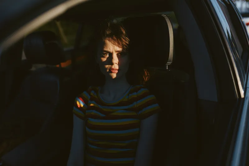 Woman sitting inside a car, striped shirt under warm retro film color, soft shadows shaping mood, intimate portrait of quiet moments behind the wheel.