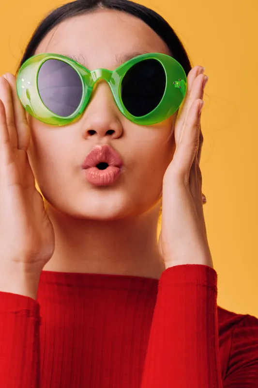 Woman wearing oversized green sunglasses and red clothing with hands near face, posing on yellow background for fashion, style and summer mood portrait closeup of female model