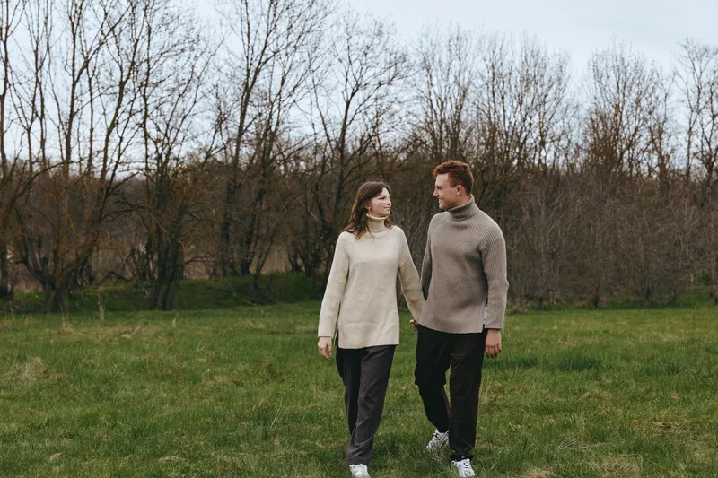 Couple outdoors in a field, walking hand in hand, cozy sweaters in neutral tones, soft sunlight, open meadow, natural colors, relaxed mood, rustic scenery during a quiet afternoon moment together