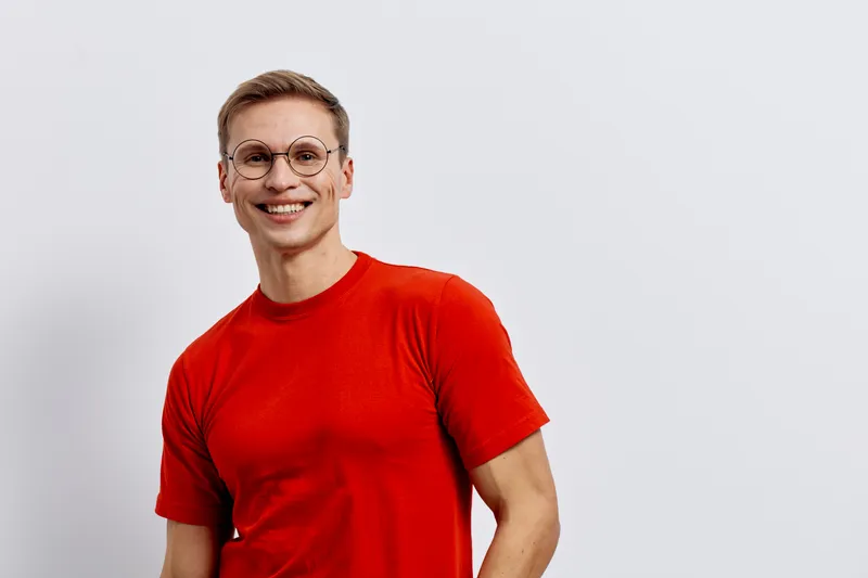 Smiling woman with short hair and glasses wearing bright red t-shirt standing against plain white background; cheerful expression, casual style, studio portrait, positive mood, confident attitude, mod