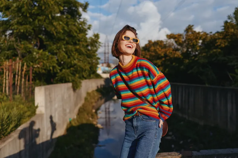 Woman in her early 20s wearing a colorful striped sweater and sunglasses, smiling while leaning on a concrete railing by an urban canal, casual outfit, travel lifestyle portrait.
