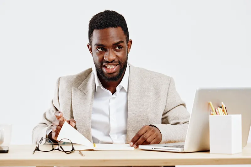 Professional young African American man in beige blazer and white shirt working at desk with laptop and glasses on a plain white background studio portrait. Business and office concept