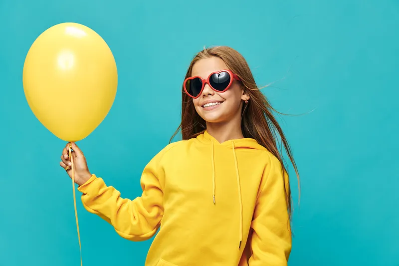 Girl with long hair and sunglasses holding yellow balloon smiling on blue background. Child, happiness and casual outfit in hoodie for playful and cheerful mood.