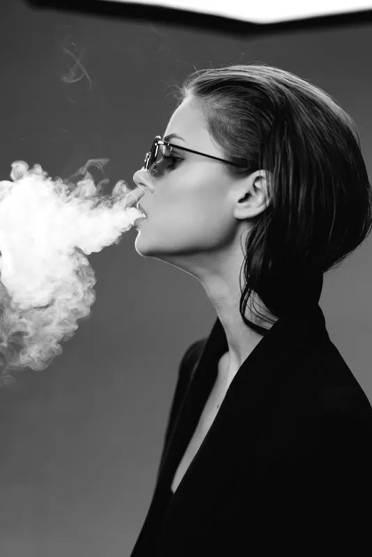 Black and white portrait of a stylish woman exhaling smoke with sunglasses and short hair, creating a moody atmosphere.