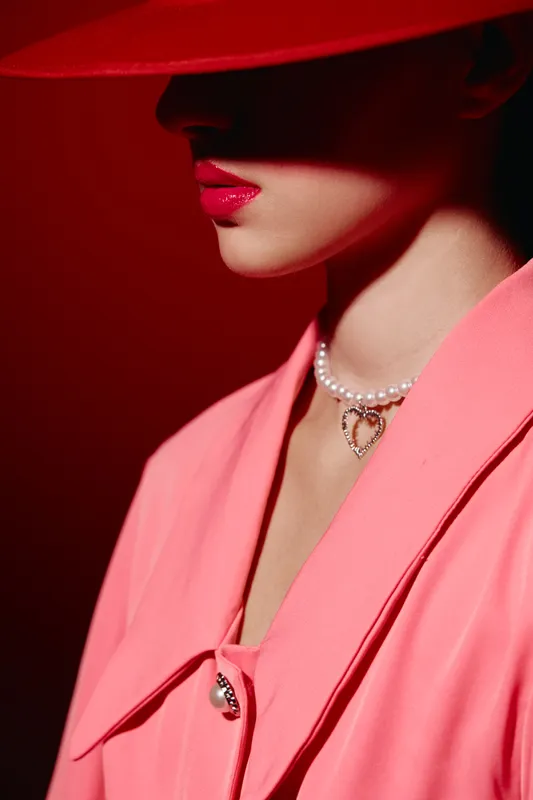 Close-up of a woman wearing a bold pink coat hat and pearl necklace with heart pendant. Dramatic lighting highlights her red lips and elegant style concept of beauty fashion creative.