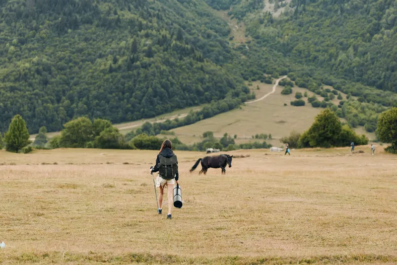 A lone traveler walks across a wide rural field toward distant hills, carrying a bag. The landscape feels calm and expansive, inviting exploration, reflection, and outdoor adventure.