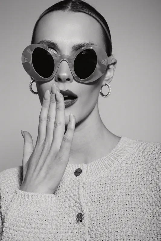 Stylish white woman with pale skin wearing round sunglasses and hoop earrings covers her mouth with hand. Fashion concept with surprised facial expression in black and white.