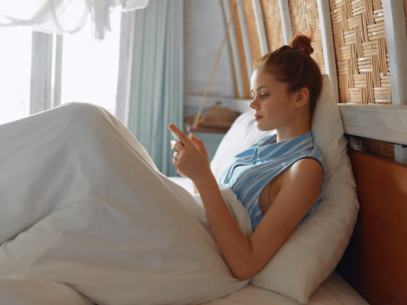 Умиротворенная женщина в постели в уютной белой рубашке и синих пижамных штанах проверяет телефон с расслабленным выражением лица