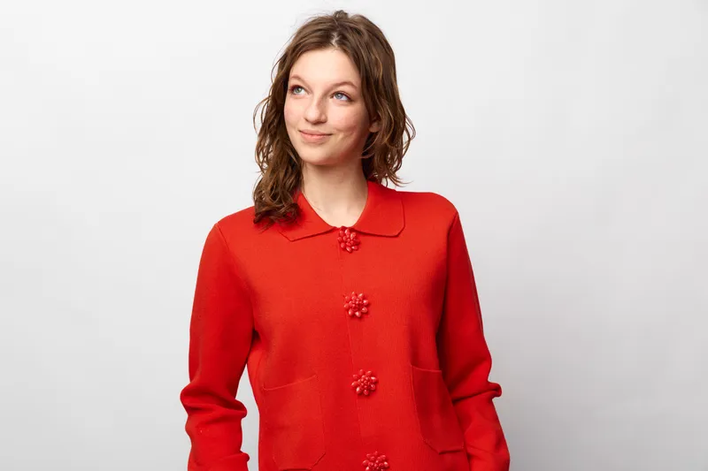 Young woman in red sweater looks sideways with slight smile on white background. Casual style, natural expression, and relaxed mood in studio portrait photography.