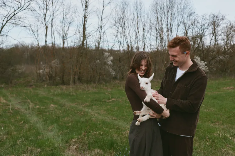 A couple outdoors in a rural meadow tenderly holds a white goat kid, sharing a joyful moment, smiling at the playful animal as gentle light and open space create a warm, candid lifestyle scene.