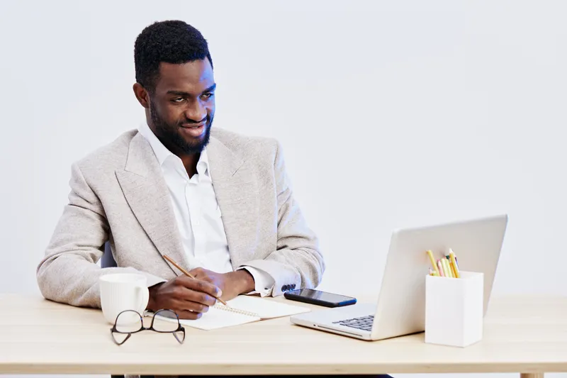 Professional smiling man in beige blazer working at desk with laptop and smartphone, wearing glasses and white shirt, isolated on plain light background, business, office, technology concept.