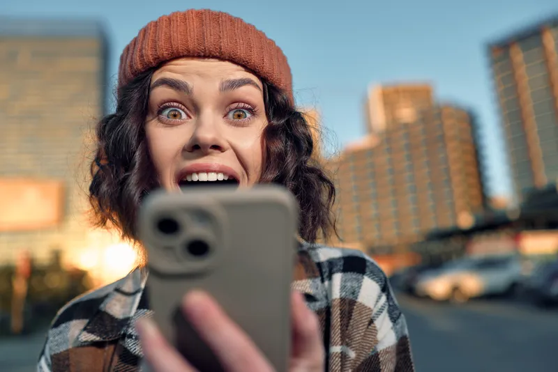 woman phone surprise city beanie selfie. Candid urban portrait of a woman staring at her phone with joyful surprise during golden hour glow, authenticity and emotional storytelling in mindful living.