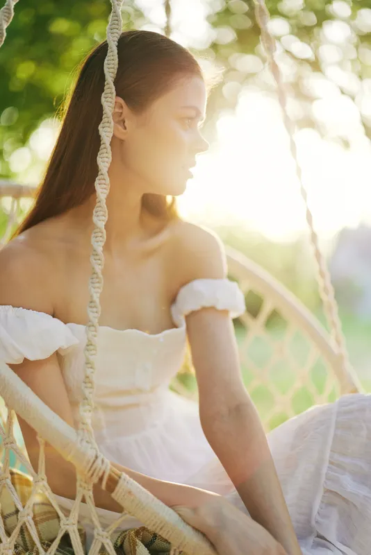 Young woman in white dress sitting on a swing, bathed in golden sunlight, conveying joy and tranquility in a lush green garden setting