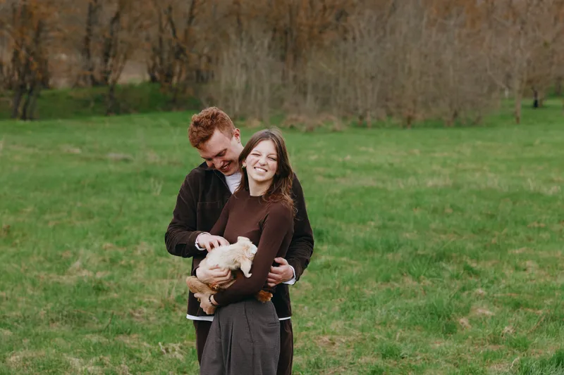 Young couple embraces in a green field while holding a small goat, sharing a joyful moment in casual autumn clothes with natural background and relaxed expressions.