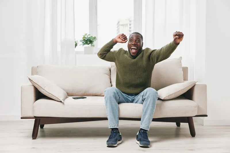 Excited young African American man celebrating at home, wearing a green sweater and jeans, showcasing joy on a bright living room sofa