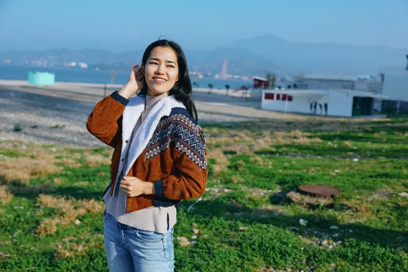 Smiling woman outdoors in a knit jacket and sweater standing in a green field with distant hills and blue sky capturing a casual outdoor fashion and nature moment