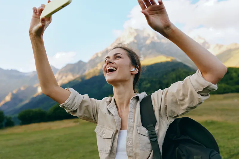 A happy young woman wearing earbuds takes a selfie outdoors in a mountainous landscape with green fields and blue sky, enjoying lifestyle and nature adventure.