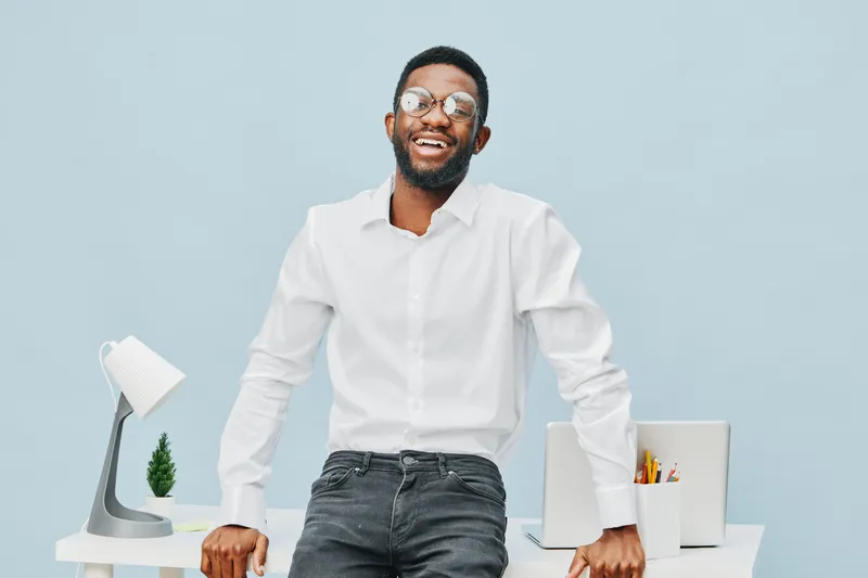 Smiling happy young man with glasses wearing white shirt standing in modern office with desk and laptop, natural light, casual style, cheerful mood, workspace setting, light blue background. People li
