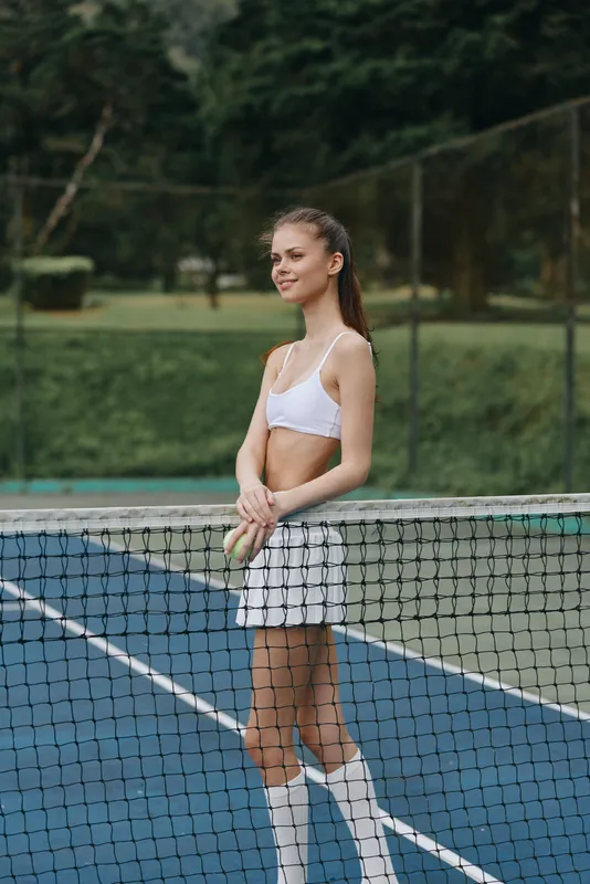 Young female tennis player in white sportswear smiling on the court, showcasing athleticism, confidence, and a vibrant outdoor background
