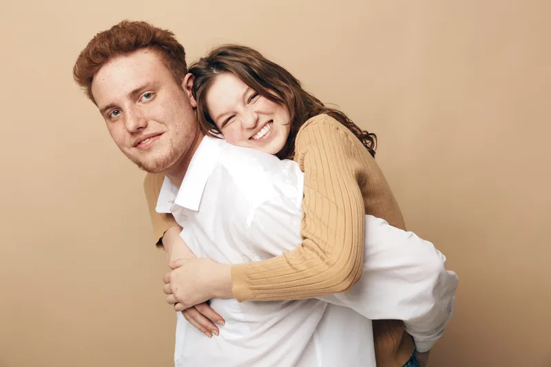 Smiling young woman in beige sweater hugs happy redhead man in white shirt on beige background. Concept of friendship, love, affection, and joyful relationship between couple or friends.