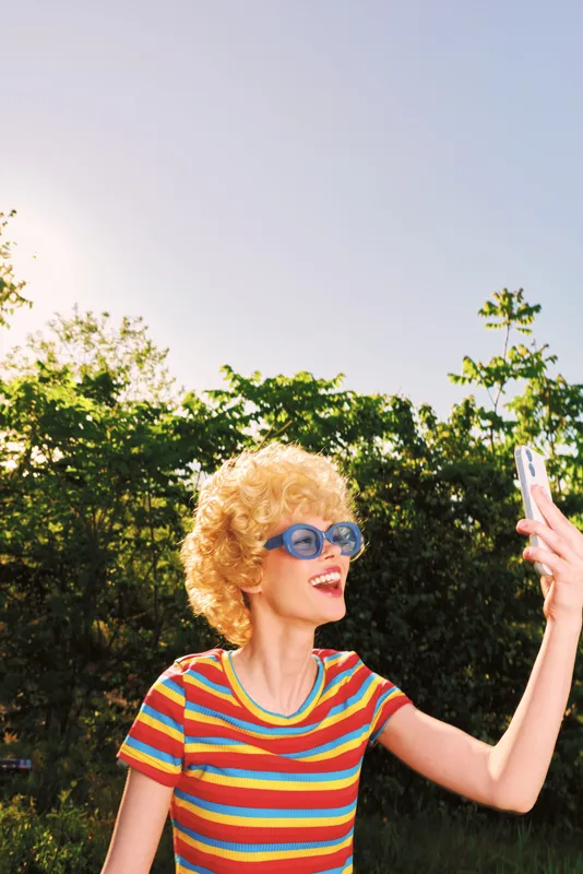 Joyful woman with curly blond hair in a striped shirt and blue sunglasses selfies outdoors, capturing a sunny, colorful moment in a park with vibrant retro tones