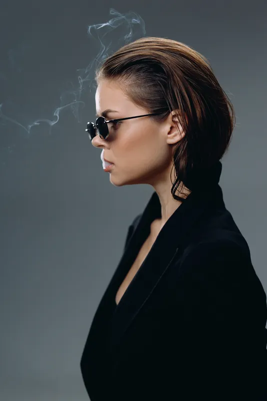 Stylish woman with short hair smoking, wearing sunglasses and dark clothing in a moody studio setting.