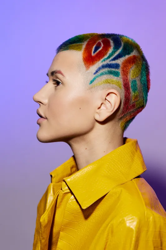 Woman with colorful short hair art and shaved patterns wearing a bright yellow jacket against a purple background in a creative style.