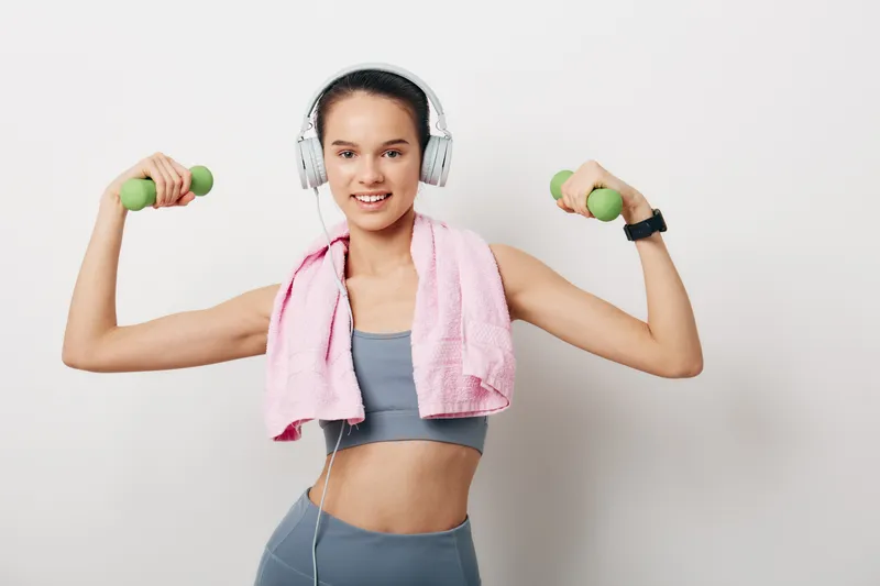 Young woman in fitness sportswear holds light dumbbells, wearing headphones and a pink towel draped over her shoulders, smiling confidently against a simple white background.
