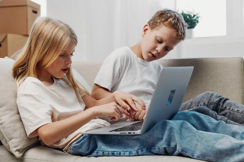 Two children sitting on a sofa using a laptop together, focused on screen in a bright living room. Concept of education, technology, and sibling interaction.