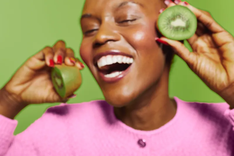 Happy African woman with dark skin smiling widely holds fresh kiwi fruit halves near face. Bright green background and pink sweater create cheerful and fresh mood. Concept of healthy lifestyle.