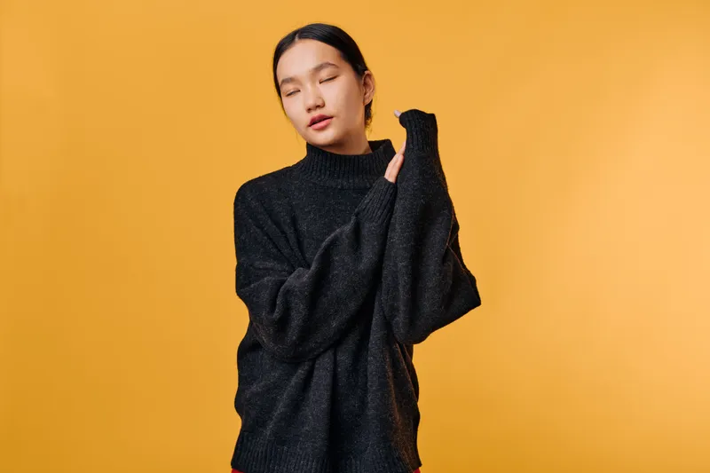 Young woman with eyes closed wearing black turtleneck sweater standing against yellow background. Portrait of serene female model in cozy warm clothing with calm expression and relaxed pose.