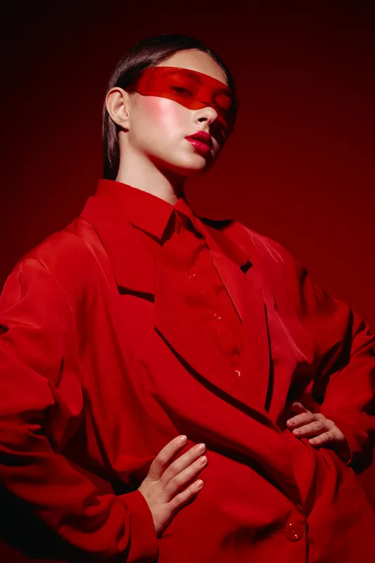 Young woman in oversized red jacket and red collar shirt poses confidently under red lighting. She wears translucent red visor glasses. Concept of creative beauty and fashion art.