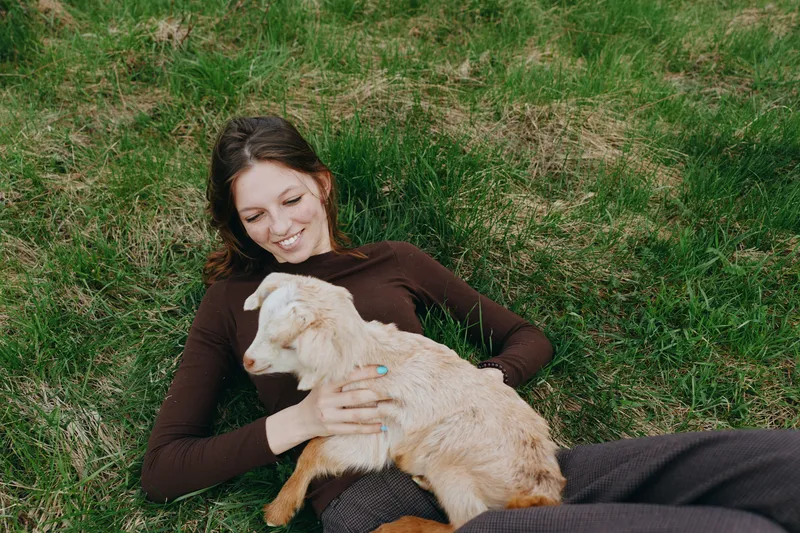 A woman lies on grass outdoors, smiling as a gentle goat kid rests in her lap. A candid lifestyle moment highlighting bonding, calm, and rural companionship.